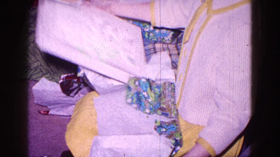 The child raises a pale sheet of wrapping or tissue as the opened floral paper rests across the lap and carpet.