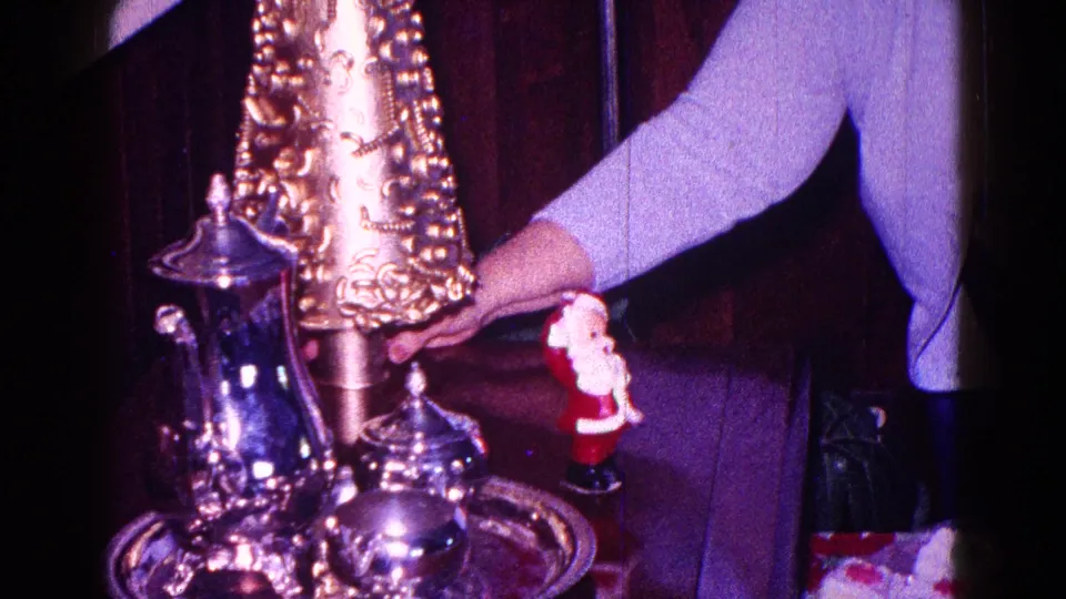 1968 Minnesota home-movie frame of a woman adjusting a gold Christmas decoration near a Santa figurine and silver teapot or coffee pot.