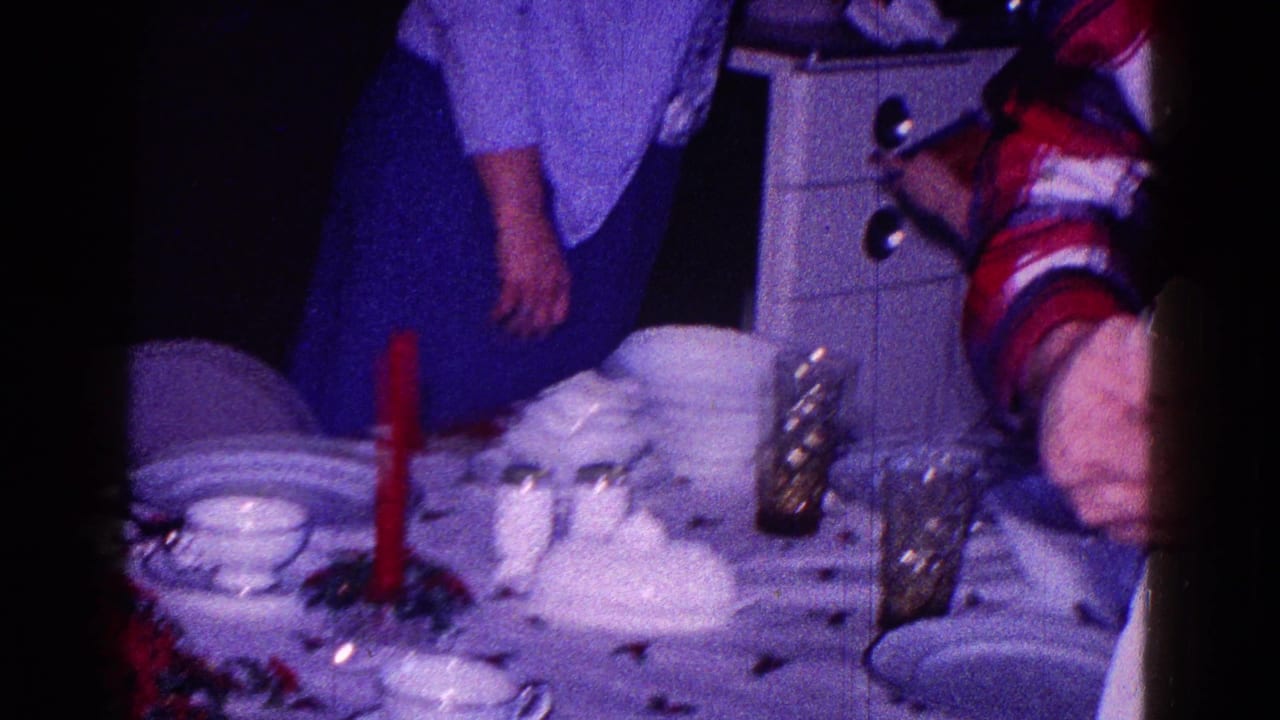 Dining table set with white dishes, a red candle, and floral holiday decoration as adults stand nearby.
