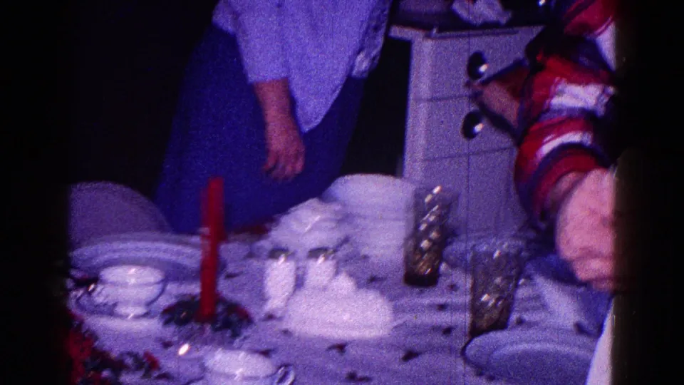 1968 Minnesota home-movie frame showing a festive dinner table with white china, a red candle, and adults partly visible nearby.