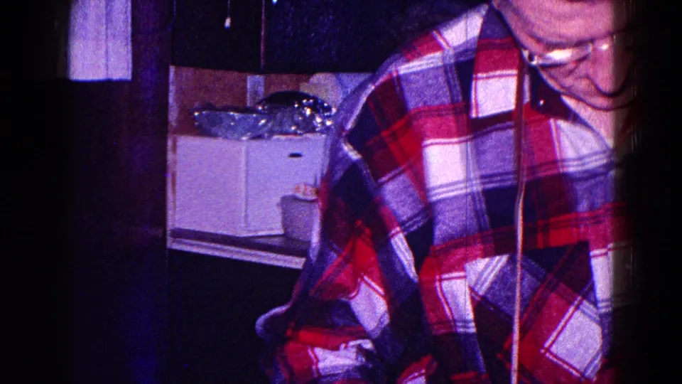 Late-1960s Minnesota home interior showing a man in a red plaid shirt near the camera, with a kitchen counter in the background.