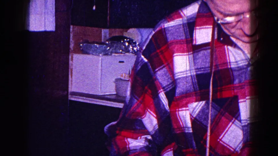 1968 Minnesota home-movie frame showing a man in a red, white, and blue plaid shirt inside a small kitchen.