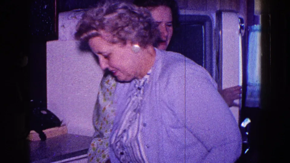 1968 Minnesota home-movie frame showing a woman in a light cardigan near a refrigerator, with another adult partly visible behind her.