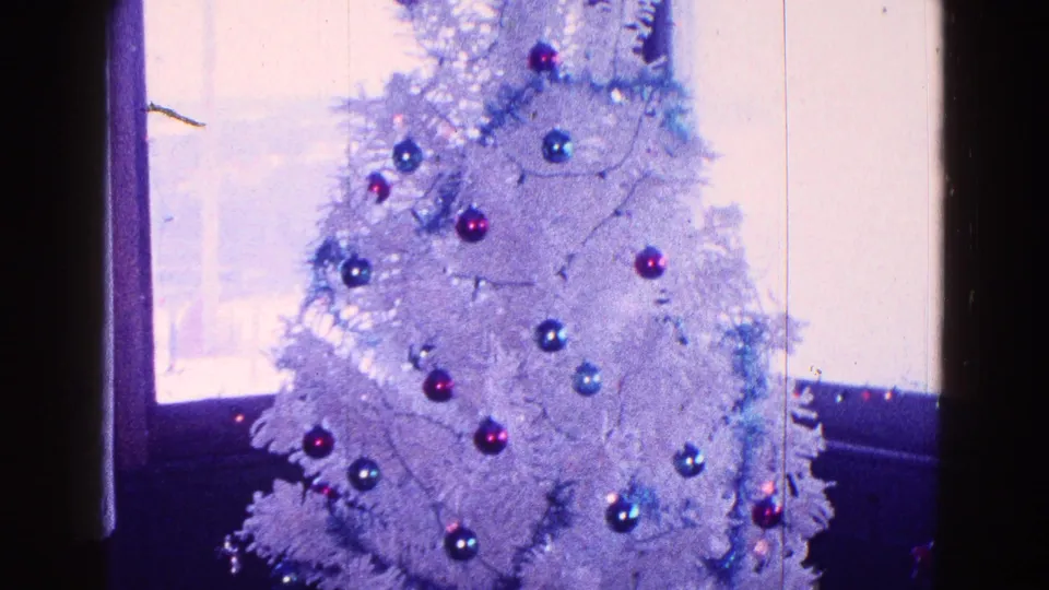 Upper portion of a white artificial Christmas tree in a 1968 Minnesota home movie, with red and blue ornaments near window light.