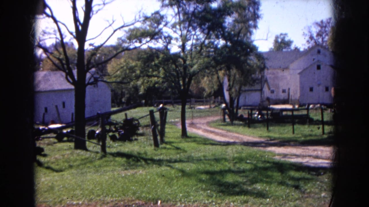 1968 Wauconda, Illinois home-movie frame showing white farm buildings, a curving dirt lane, trees, fencing, and equipment.