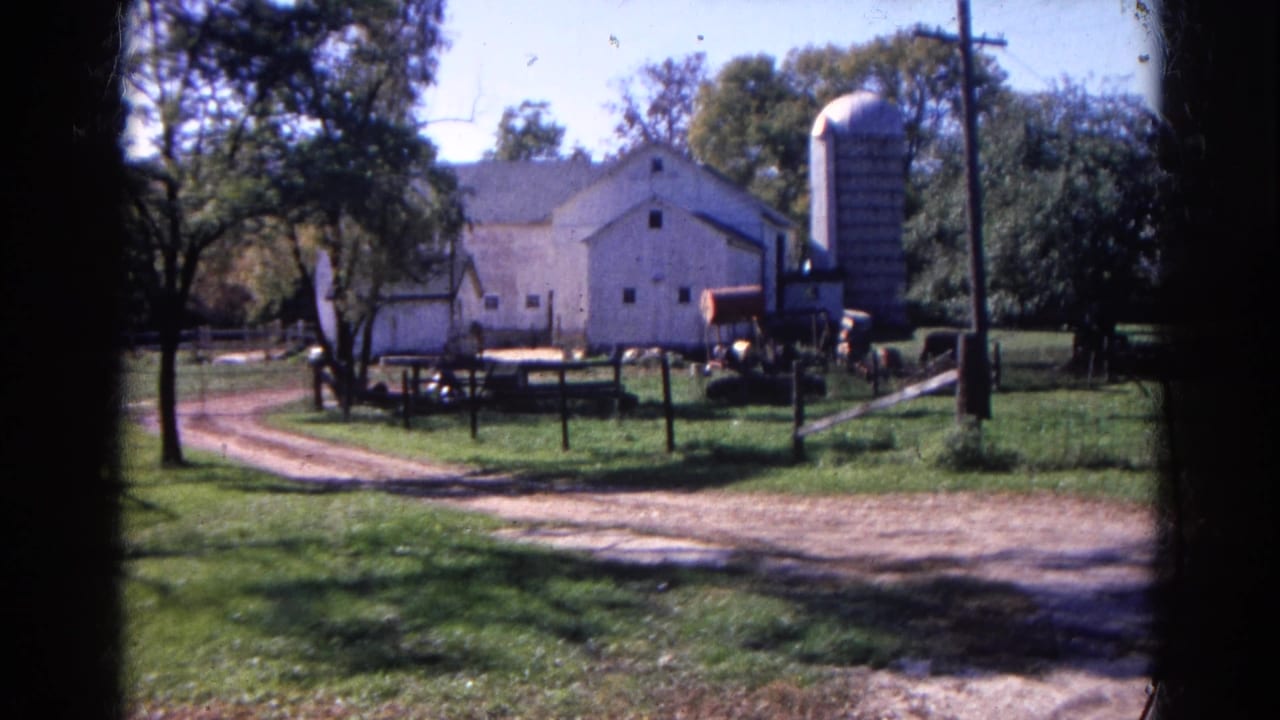 White barn and silo across a fenced farmyard with a dirt driveway and trees.