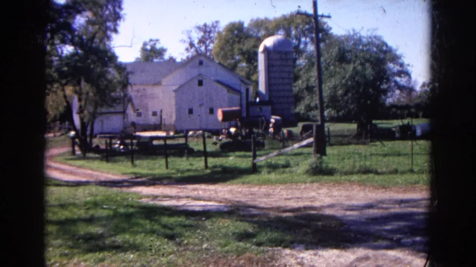 The 1968 Wauconda farm remains framed from outside, showing the white barn, upright silo, fenced grass, dirt drive, trees, and poles.