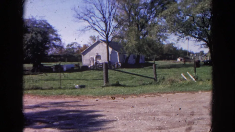1968 Wauconda farmyard view with white building, trees, fence posts, grassy ground, dirt road, and distant small structures.