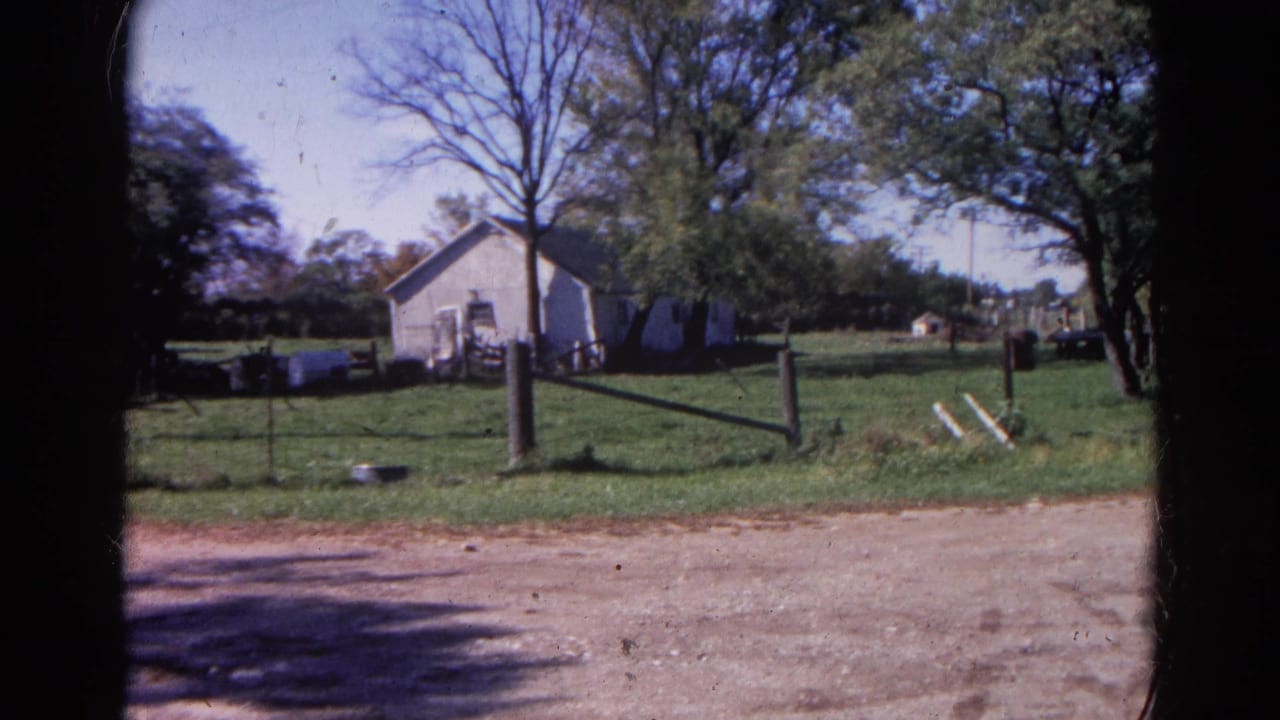 White farm building behind a fence with grass, trees, and a gravel foreground.