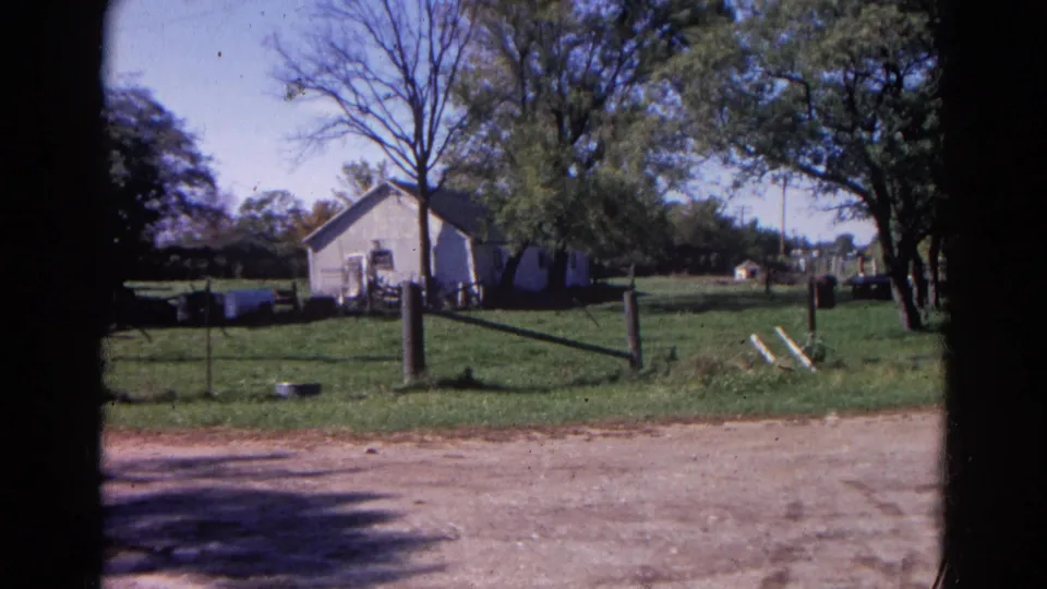 Steady 8mm view of a rural Wauconda farmyard with a white building, wooden fence, grass, trees, and animals behind the fence.