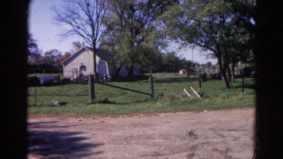1968 rural Illinois home-movie frame with a white farm building, fence posts, green yard, trees, and dark 8mm frame edges.