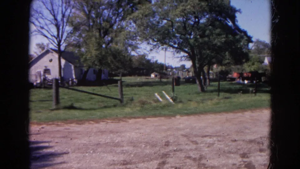 Wauconda 8mm frame showing the same fenced rural yard, white building, trees, and a red vehicle or equipment partly visible at right.
