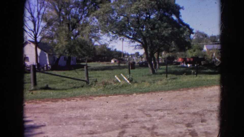 1968 Wauconda, Illinois home-movie frame showing a grassy rural yard, wooden fencing, trees, and houses beyond a dirt foreground.