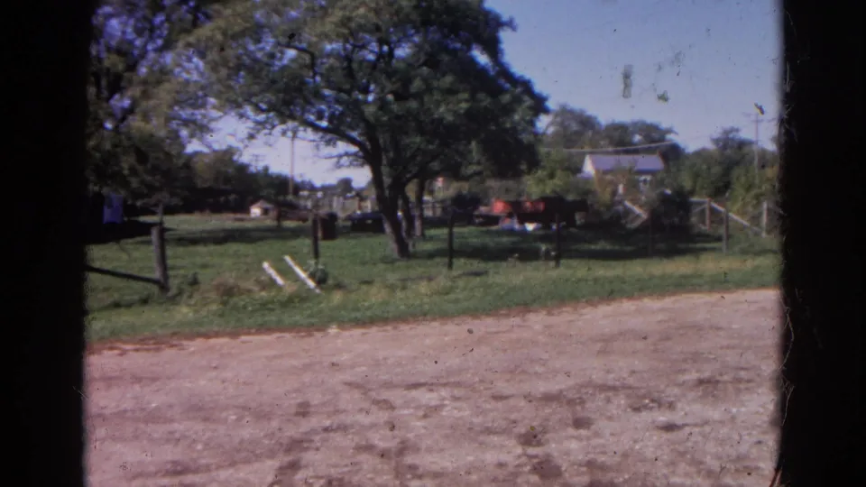 1968 Wauconda, Illinois frame showing leaning white boards near a fence, trees, grass, and neighboring structures in soft 8mm color.
