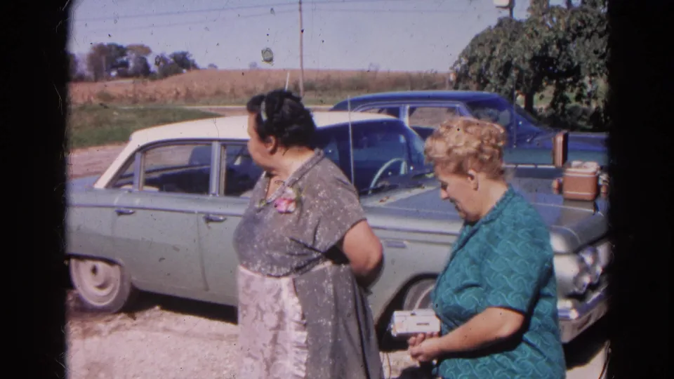 Two women in 1968 Wauconda stand beside a light green sedan, with a blue car and fields behind them.