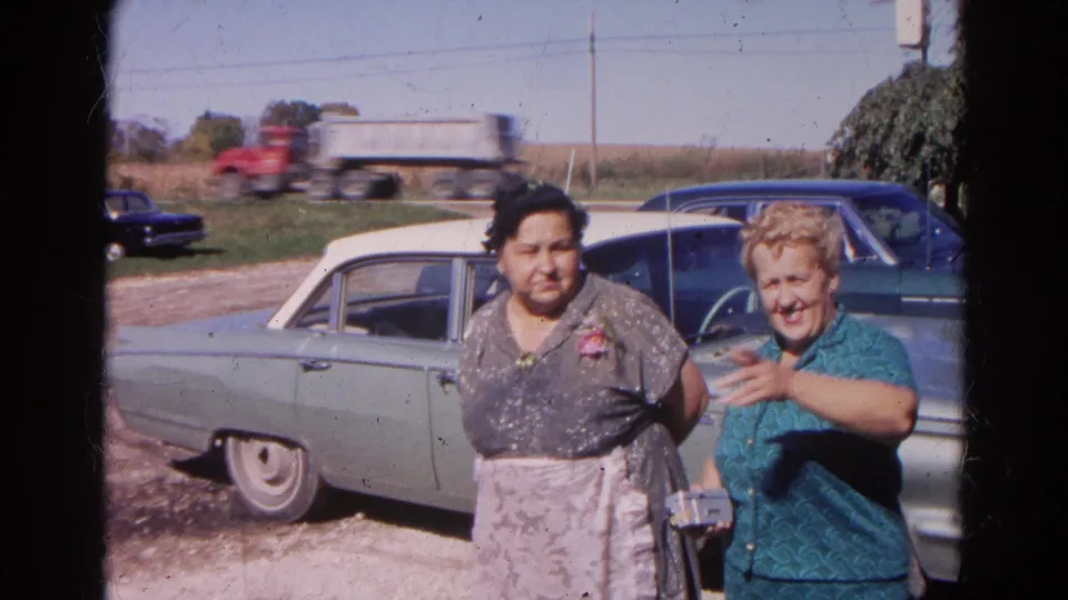 In a 1968 Wauconda farm setting, two women face the lens by a sedan as one holds a small camera and a truck passes.