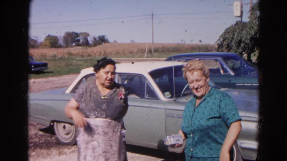 Two women in a 1968 Wauconda home movie smile and gesture beside a light green sedan on a rural roadside.