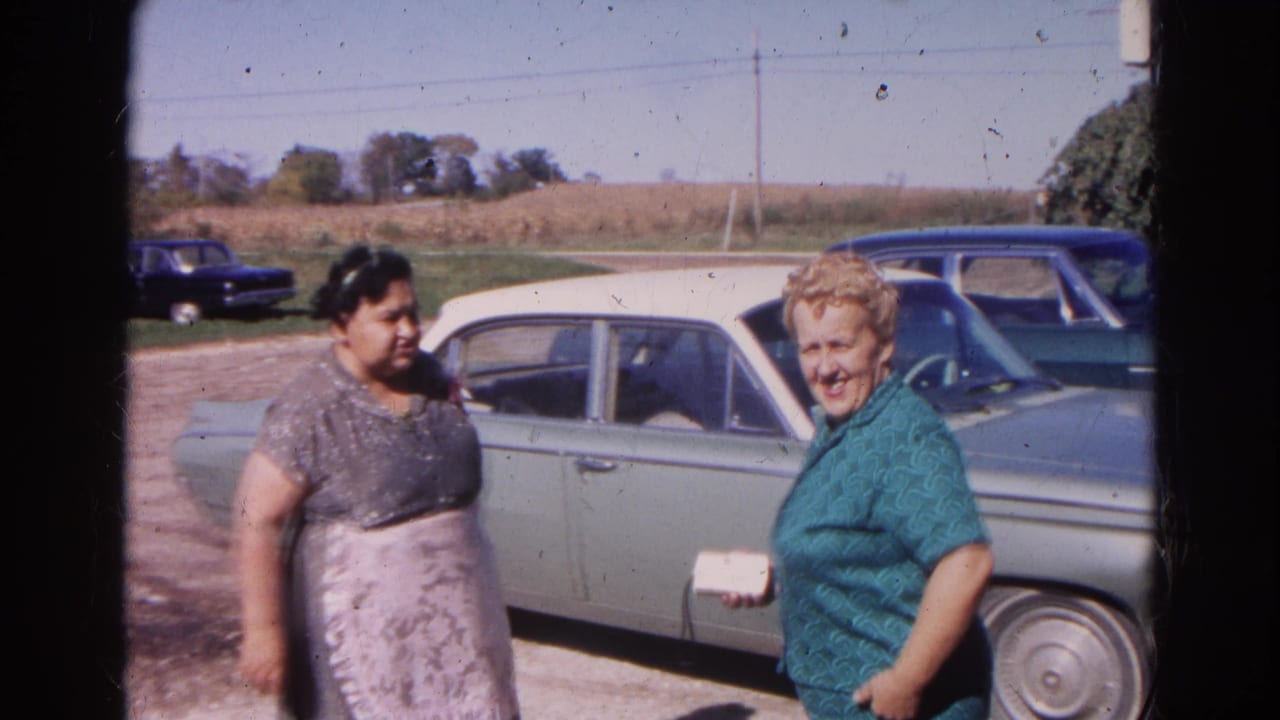 Two women stand beside a pale green and white sedan in a rural yard.