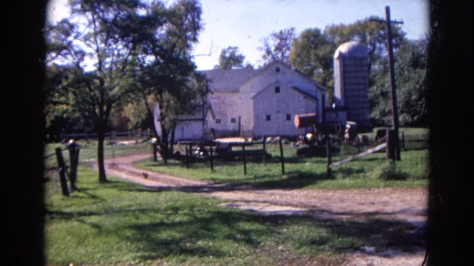Wauconda, Illinois farmyard in 1968 with white barn, silo, fenced pasture, trees, and dirt drive under bright sky.