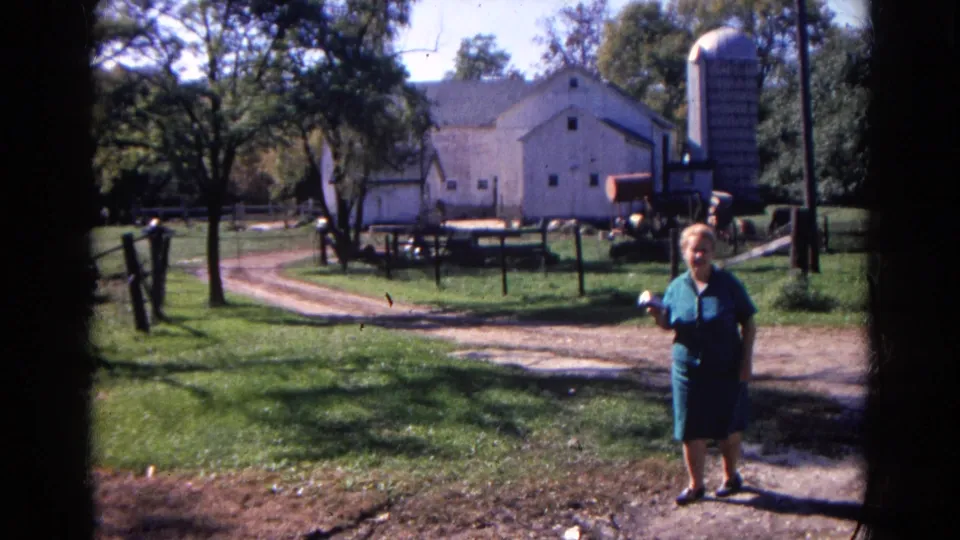An adult woman in a blue dress approaches the camera with a barn, silo, fencing and dirt lane behind her.