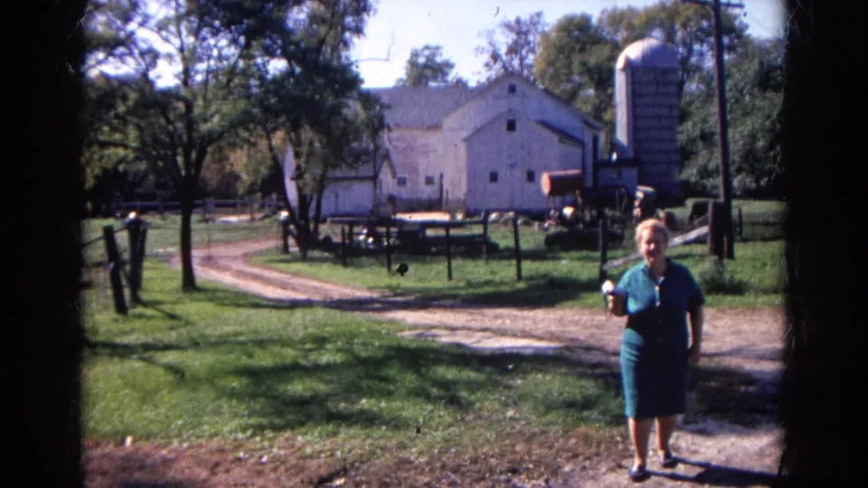 The woman in blue stands closer to the camera as the white barn, silo, trees and fenced yard remain visible behind her.