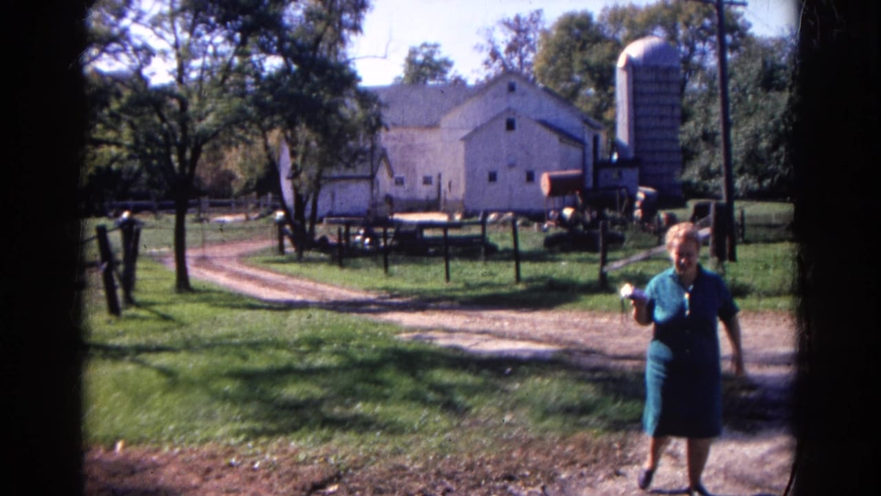 Woman in a blue dress walks toward the camera in a farmyard with a barn and silo behind her.