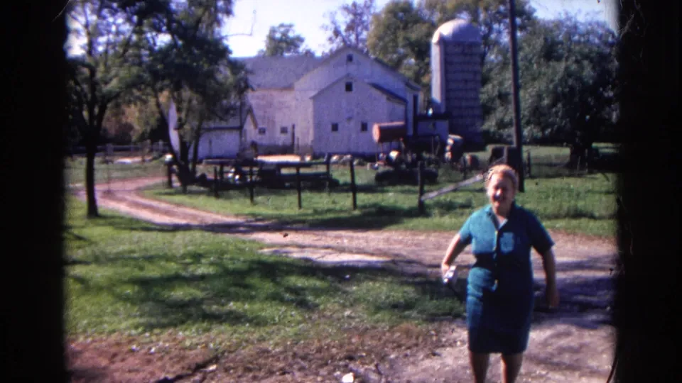 The woman continues toward the camera in the 1968 Wauconda farmyard, with the white barn, silo, fencing, grass, and trees still visible.
