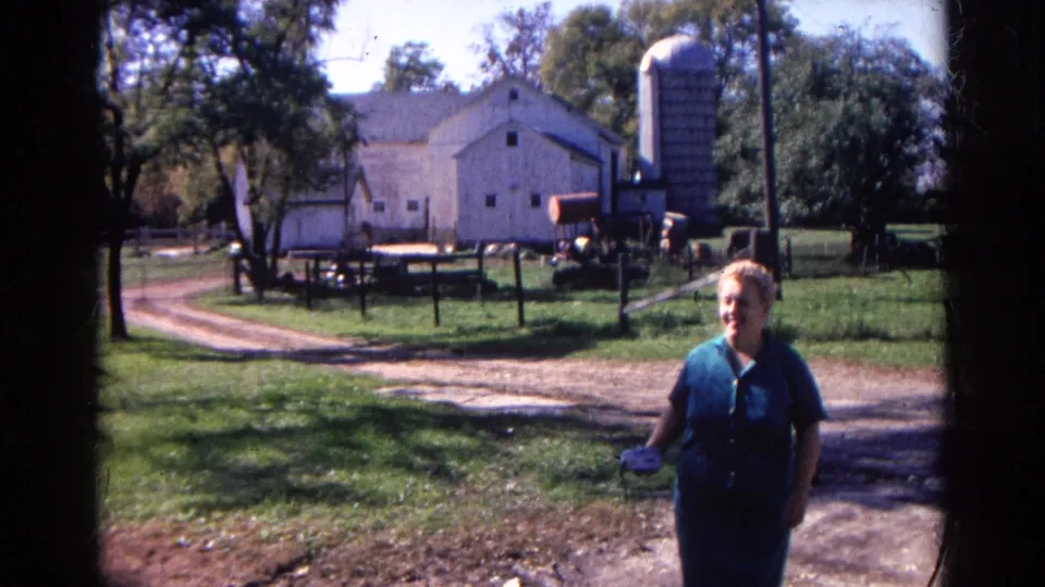 Nearer the camera, the woman in the blue dress is seen in the Wauconda farmyard with a barn, silo, fenced grass, and farm equipment behind her.