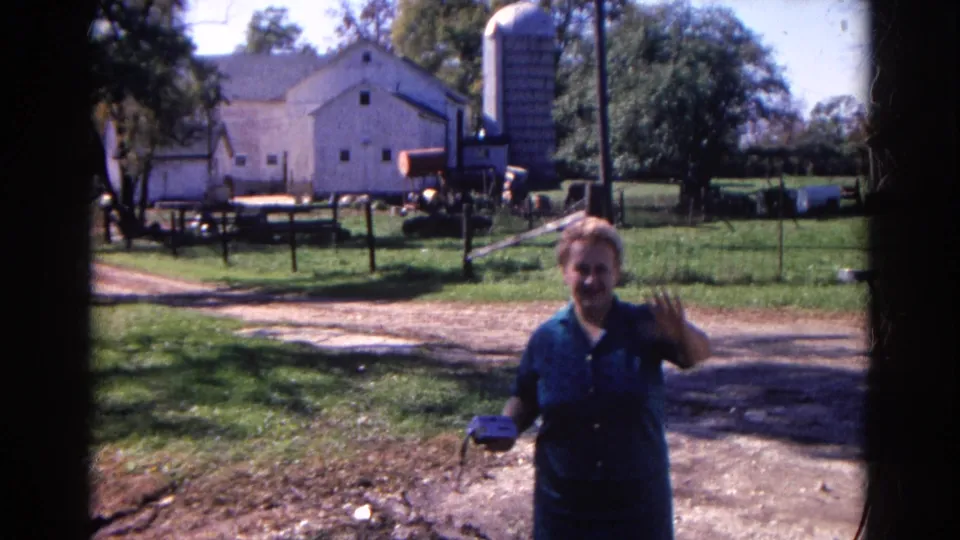 In the final frame, the woman waves near the camera in the 1968 Wauconda farmyard, with the barn, silo, dirt lane, fencing, and trees behind her.