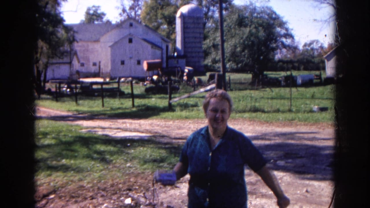 In 1968 Wauconda, Illinois, a woman walks toward the camera on a rural farm lane with a white barn and silo behind her.