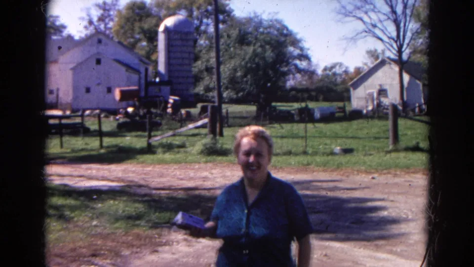 The woman continues toward the camera on the rural Illinois farm, framed by fencing, trees, a barn, and a silo.