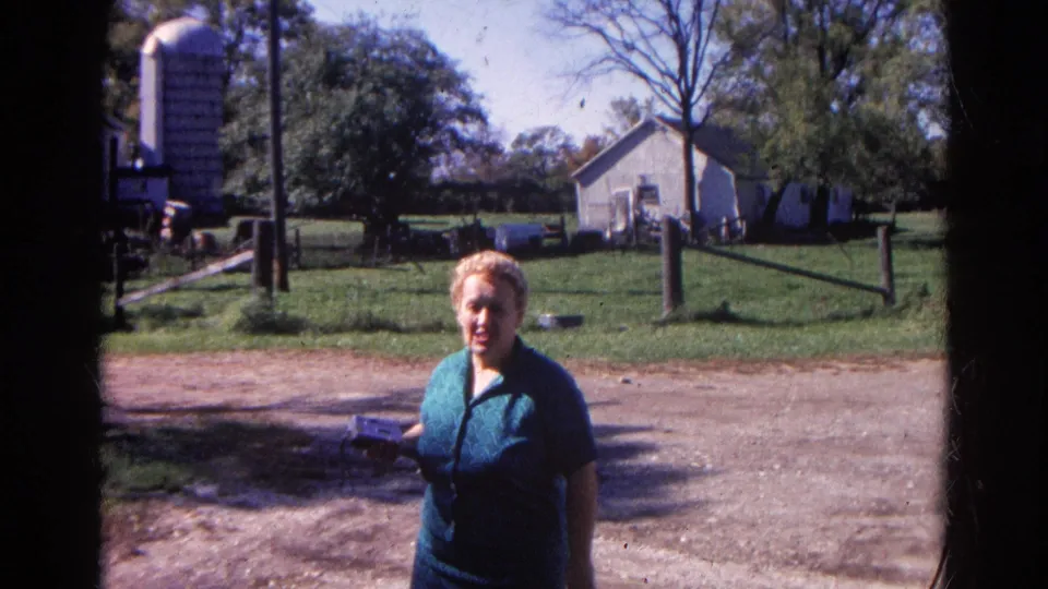 Near the end of the Wauconda farm clip, the woman stands on the lane with fenced grass, outbuildings, and trees visible behind her.