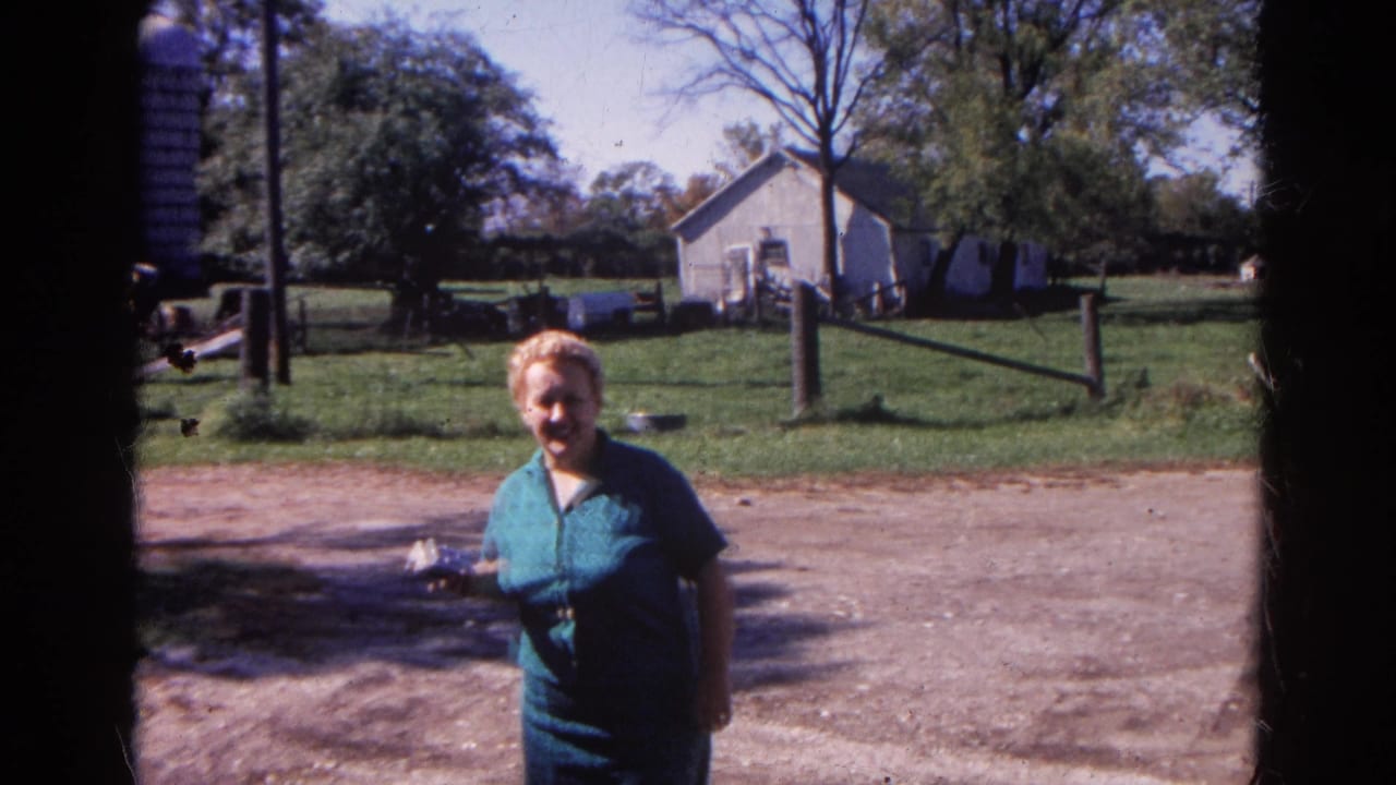 Adult woman standing in a rural farmyard with trees, fencing, and outbuildings behind her.