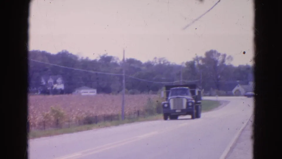 Dark dump truck approaches the camera on a two-lane rural road in 1968 Wauconda, Illinois, with cornfield and farm buildings nearby.