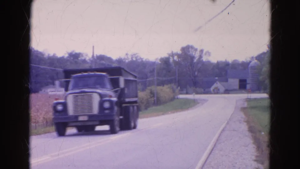 A dark dump truck passes close on a rural Wauconda road, with a barn, silo, cornfield, and pale houses in the background.