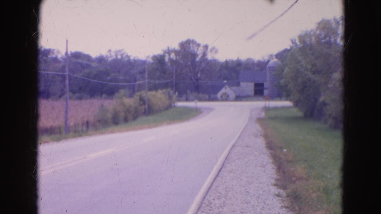 1968 Wauconda roadside view with a curving asphalt road, grassy shoulder, utility lines, trees, and distant farm buildings.