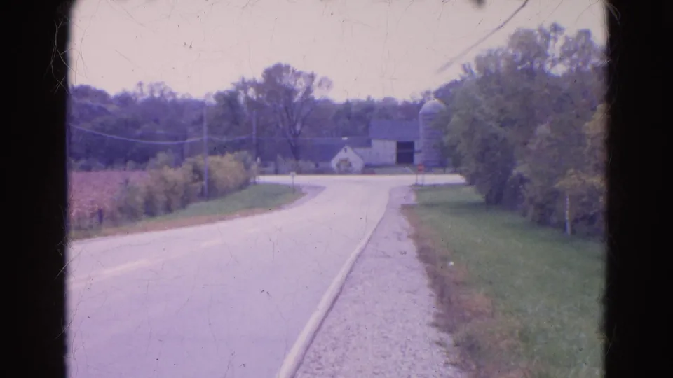 1968 Wauconda view down a rural road toward a white farm building, silo, grassy verge, fields, and tree line.