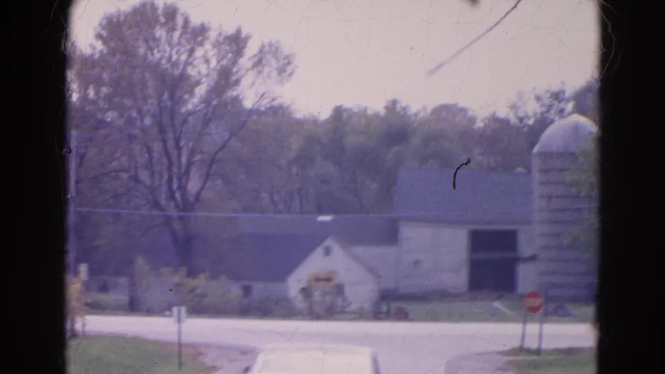 Closer 1968 Wauconda farmstead view showing a white building, silo, roadside stop sign, trees, and pale road surface.