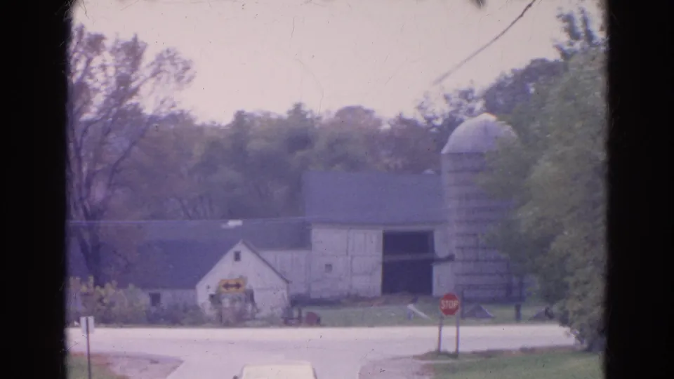 1968 Wauconda, Illinois home-movie frame showing a white sedan on a rural road near a barn, silo, trees, and stop sign.