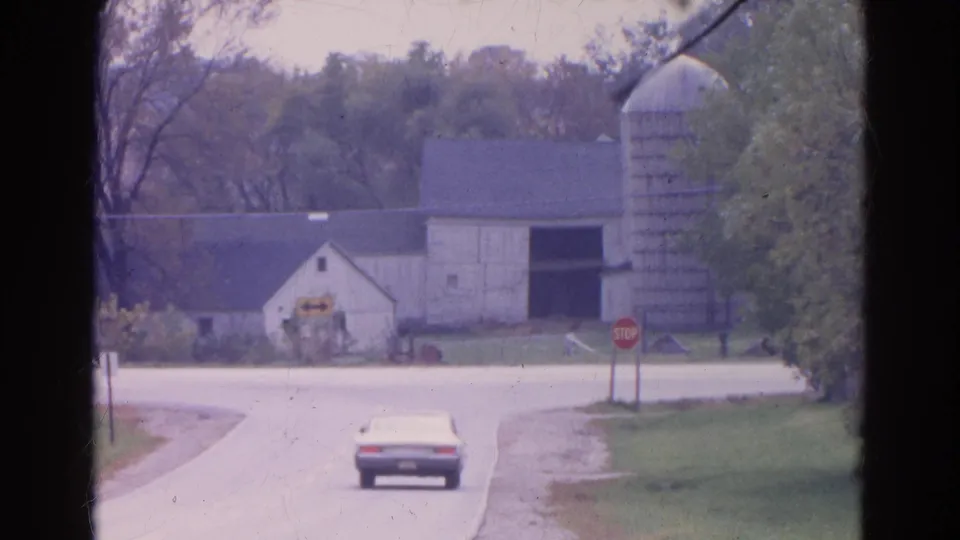 1968 Wauconda, Illinois frame with a white sedan moving away toward a farm complex, tall silo, stop sign, and wooded background.