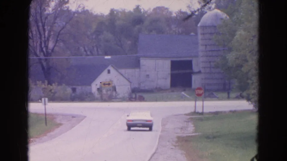 1968 Wauconda, Illinois home-movie still of a white sedan receding on a country road near a barn, silo, and red stop sign.
