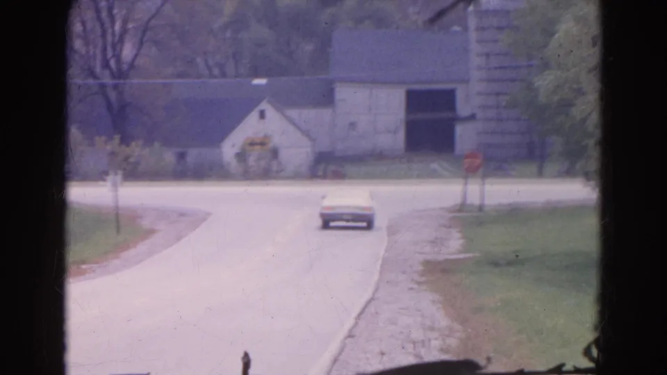 1968 Wauconda, Illinois frame showing the sedan farther from camera on a rural road with farm buildings, silo, and stop sign nearby.