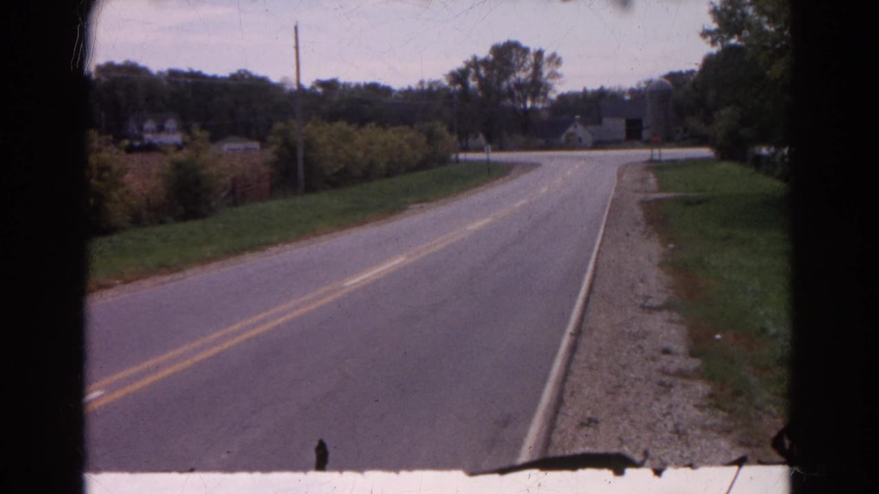 A two-lane country road curves toward distant farm buildings and trees.