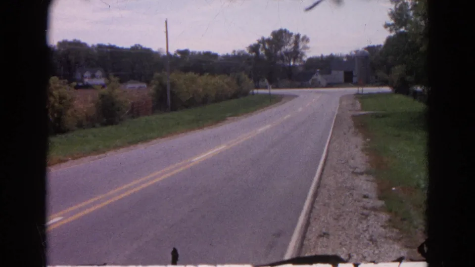 1968 Wauconda, Illinois 8mm frame of a quiet paved road curving through a rural landscape with brush, grass, distant buildings, and trees.