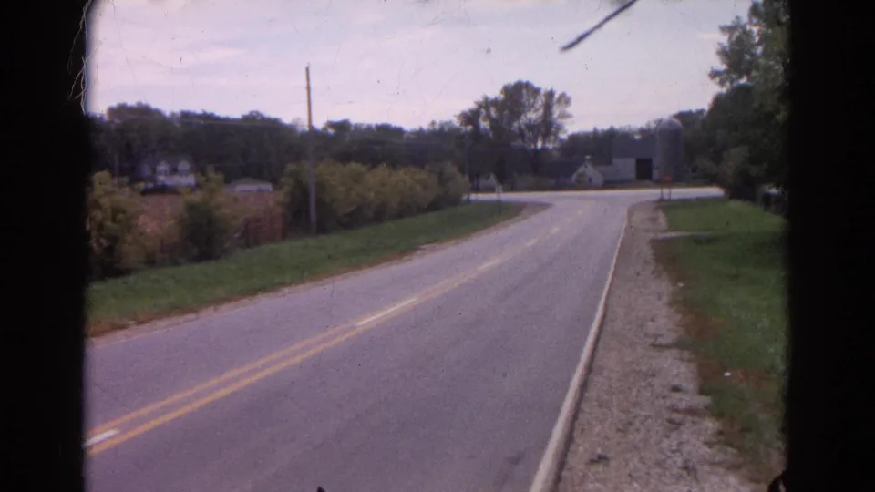 1968 Wauconda, Illinois rural road frame showing a two-lane roadway leading toward an intersection, farm buildings, trees, and a gravel shoulder.