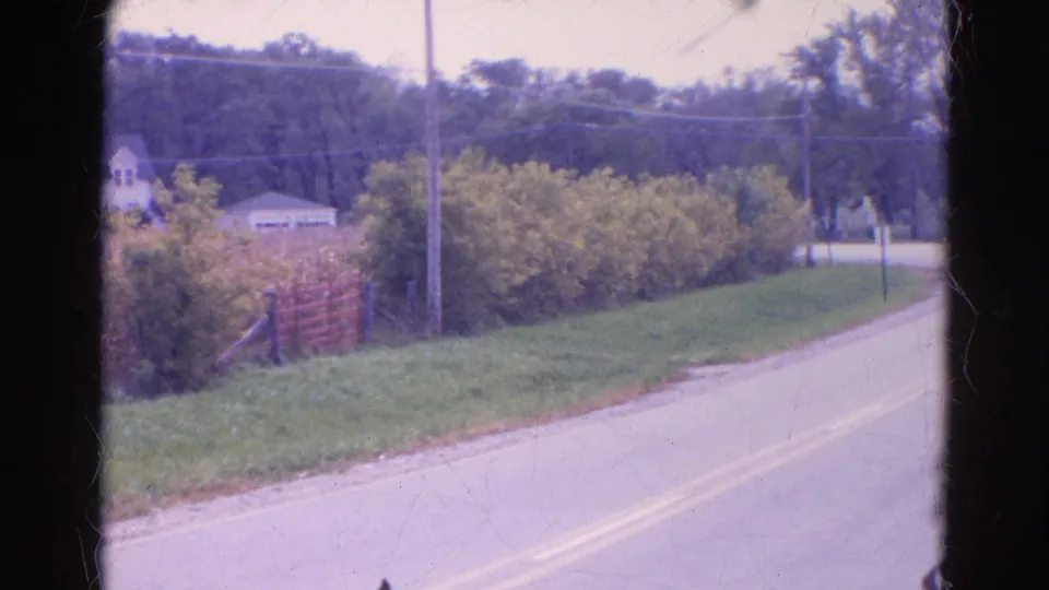 1968 Wauconda, Illinois roadside view showing a bend in the road, thick greenery, a utility pole, and small buildings near a wooded slope.