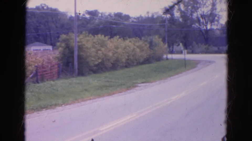 1968 Wauconda, Illinois home-movie frame of a rural road edge with yellow center lines, green shoulder, bushes, utility lines, and trees beyond.