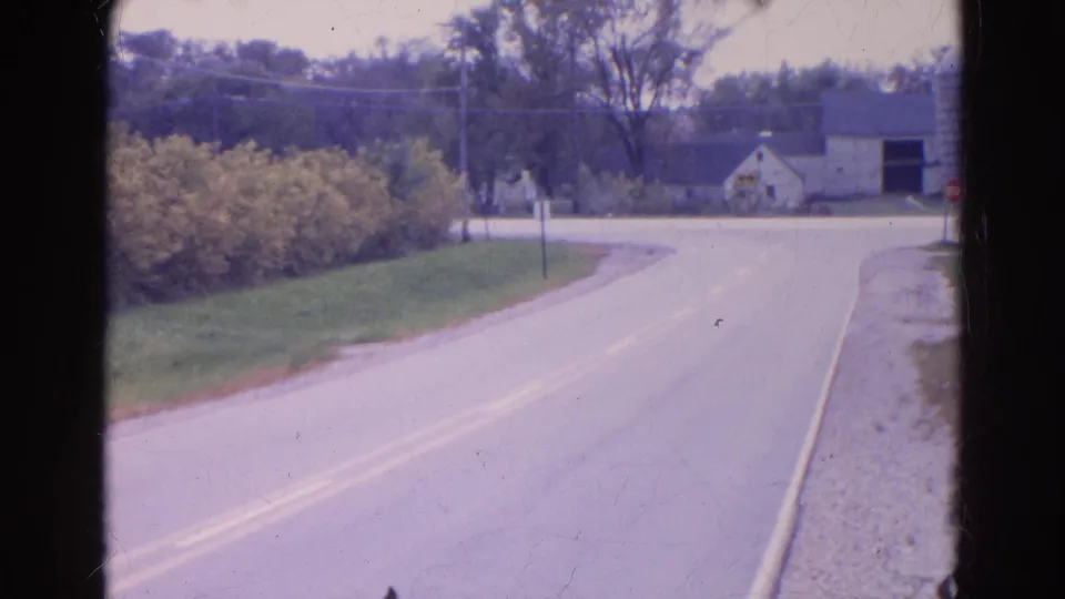 1968 Wauconda, Illinois view of a curving rural road with roadside bushes, grassy shoulder, trees, and a farm building near the right edge.