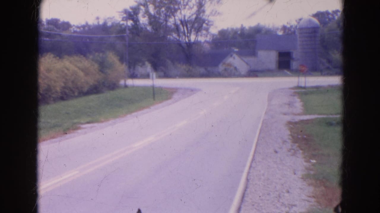 Curving paved road leading toward a farmstead and silo near a stop sign.