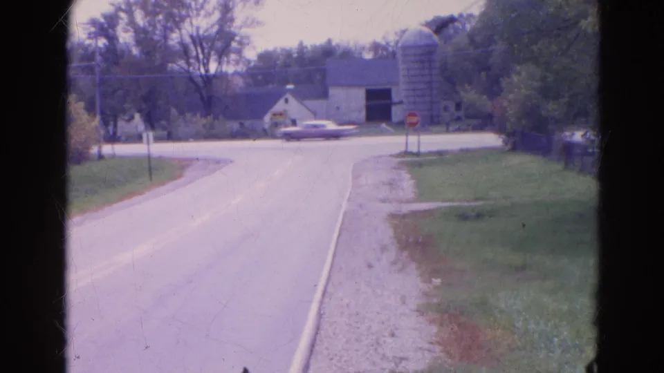 A 1968 Wauconda road scene shows a car passing across the far intersection near a farmstead, silo, stop sign, and trees.
